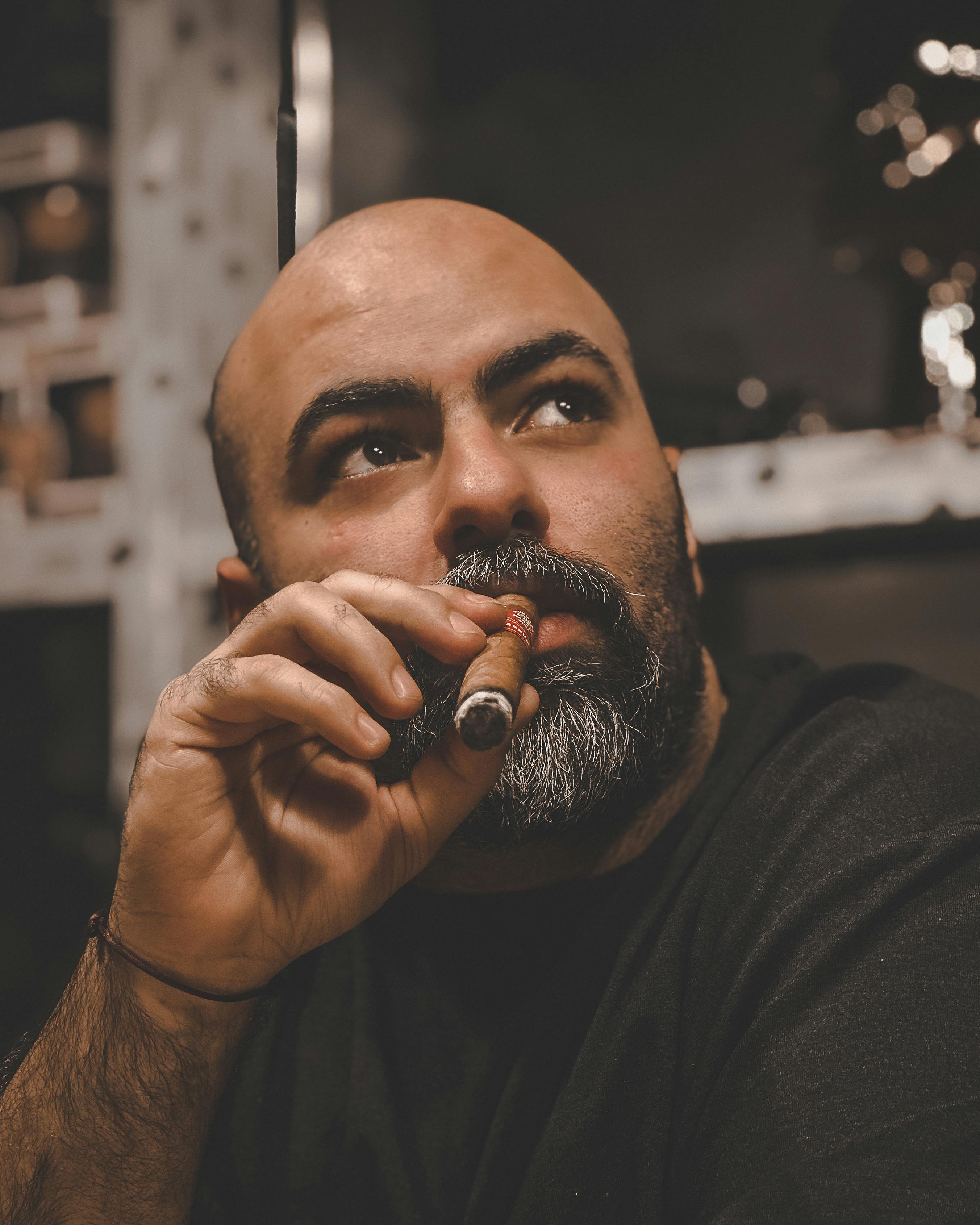 Close-up of a bald, bearded man enjoying a cigar indoors with a thoughtful expression.