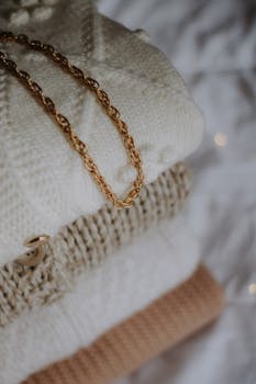 Stack of warm knit sweaters with a gold chain necklace for a cozy winter vibe.