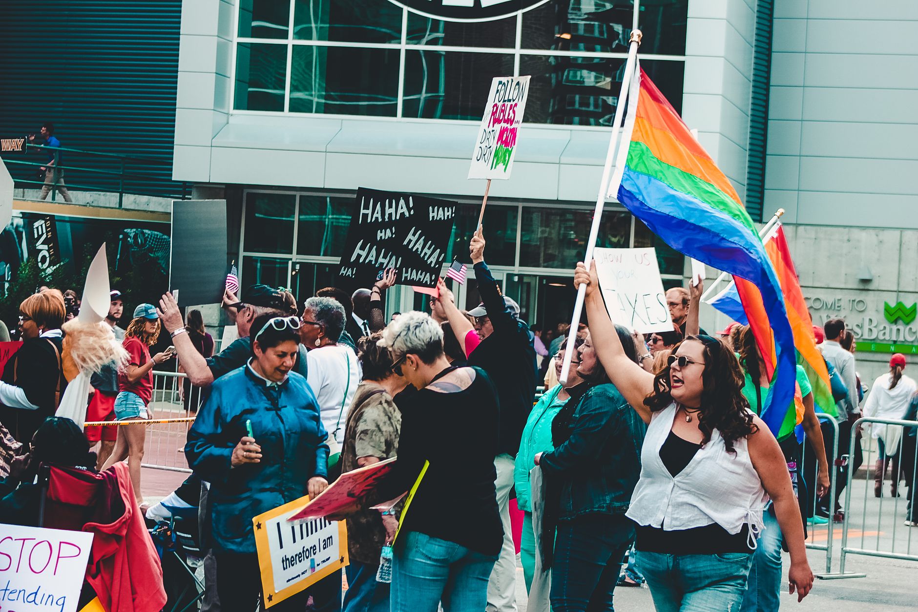 Manifestanti in una strada con cartelli e una grande bandiera arcobaleno sollevata davanti a un edificio vetrato