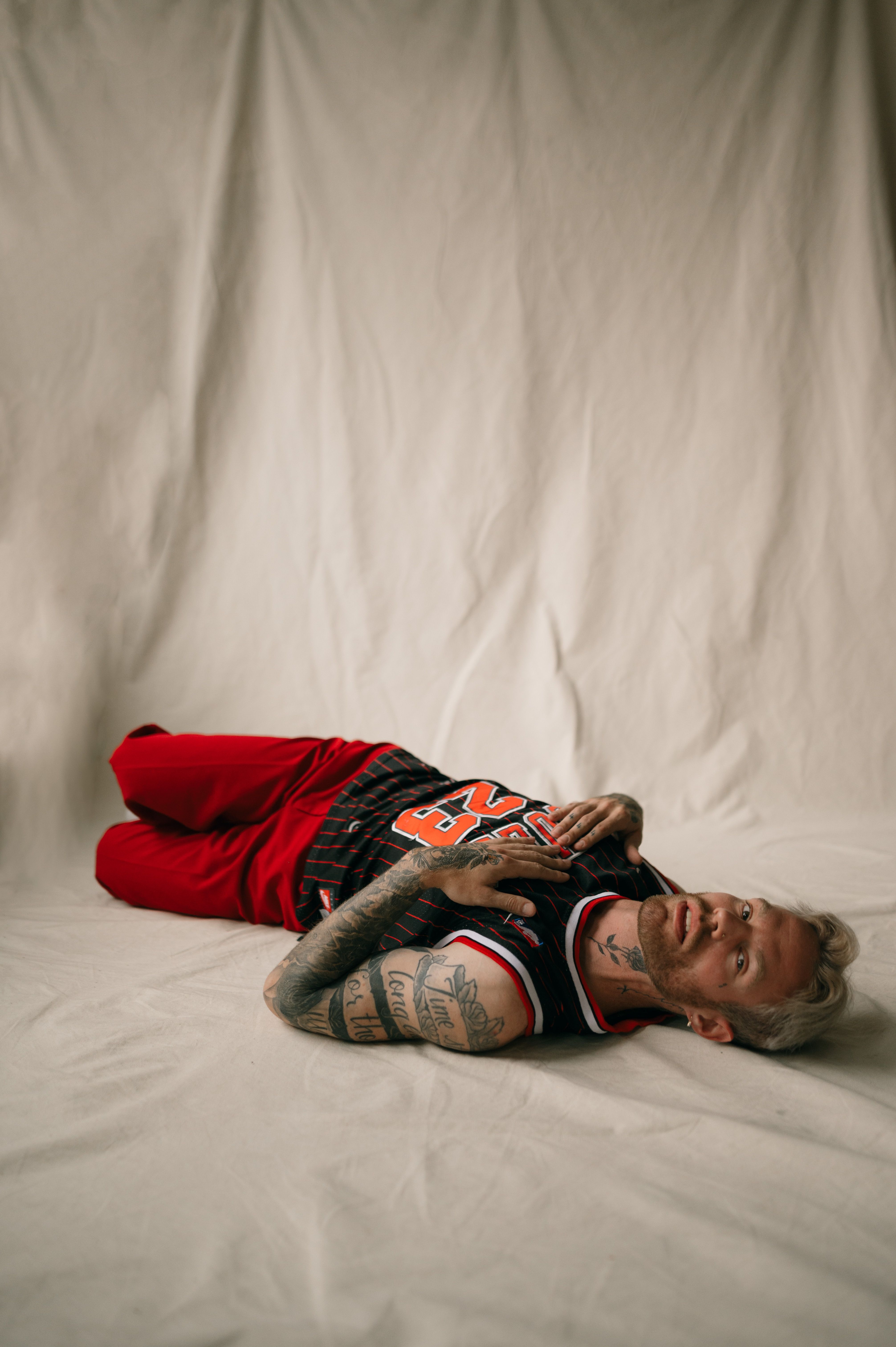 A tattooed man lies on white fabric, posing in a basketball jersey and red shorts with a neutral expression.