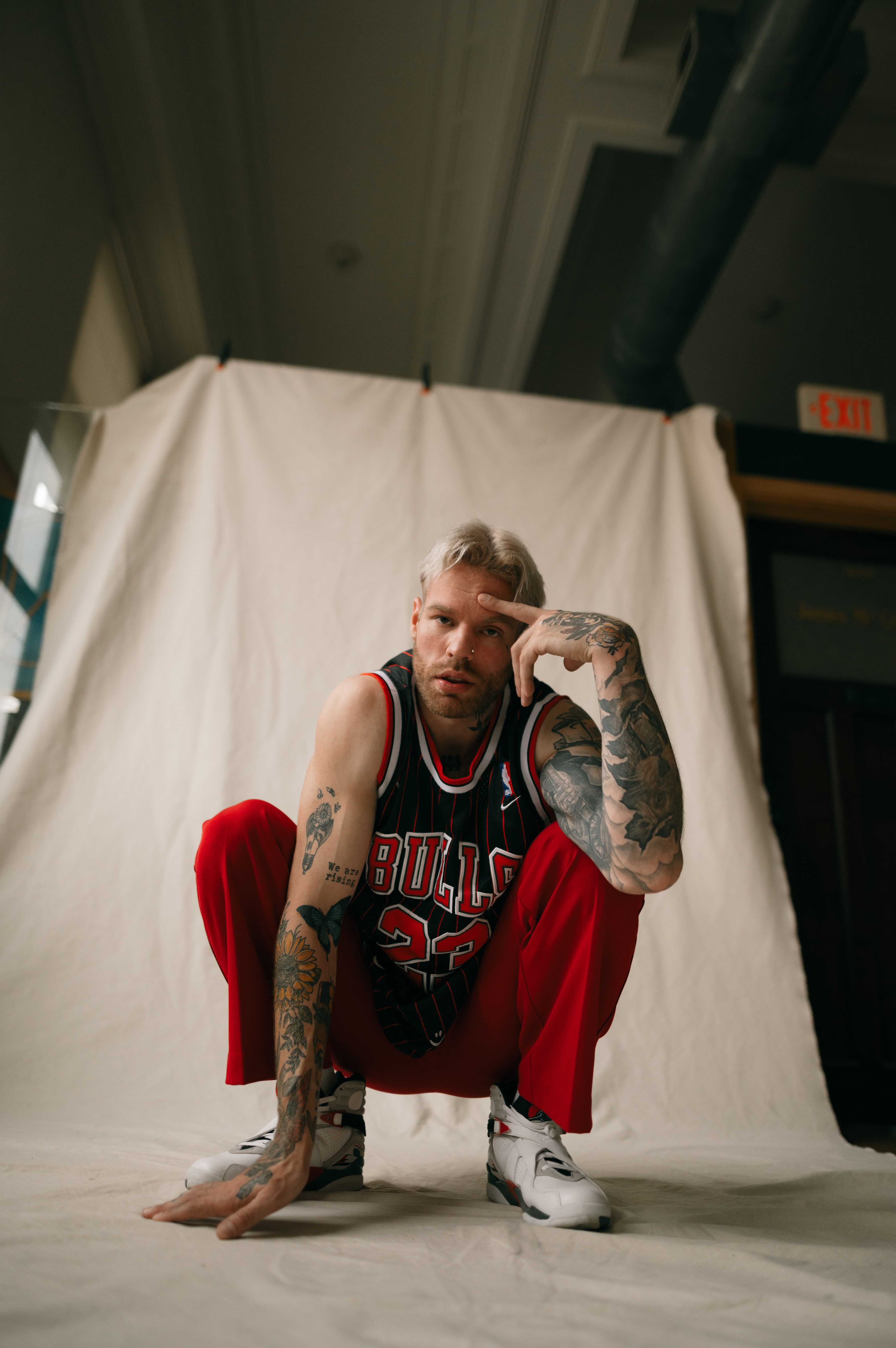 Tattooed man crouching in a basketball jersey with red pants against a white fabric backdrop.