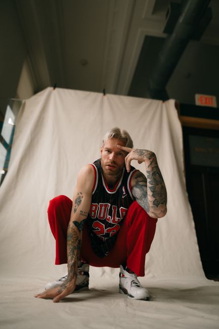 Tattooed man crouching in a basketball jersey with red pants against a white fabric backdrop.