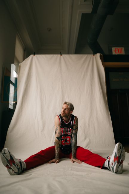 Athletic man in a basketball jersey poses indoors, showing his tattoos and stylish sneakers.