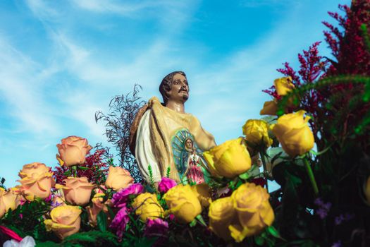 A vibrant statue of religious figure with colorful roses under a clear blue sky symbolizing faith and spirituality.