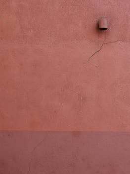 A vertical shot of a rough pink wall with a noticeable crack and small pipe.