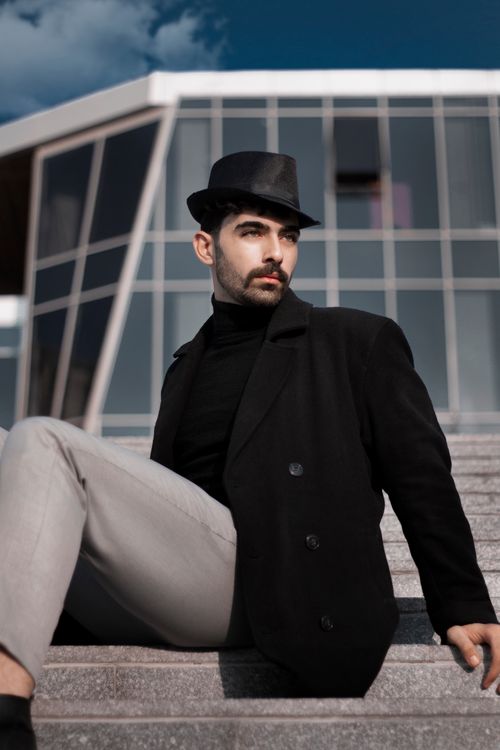 Free A fashionable adult man in a black coat and hat sitting on steps against a modern glass building. Stock Photo