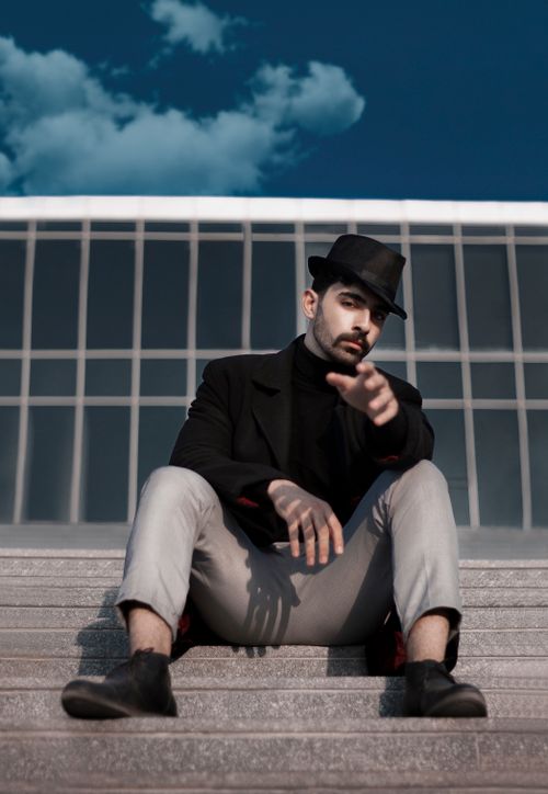 Free Stylish man in a hat sitting on stairs against a modern building with blue sky. Stock Photo