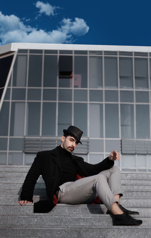 Free A fashionable man in black attire and fedora hat sits on modern city steps against a geometric building backdrop. Stock Photo
