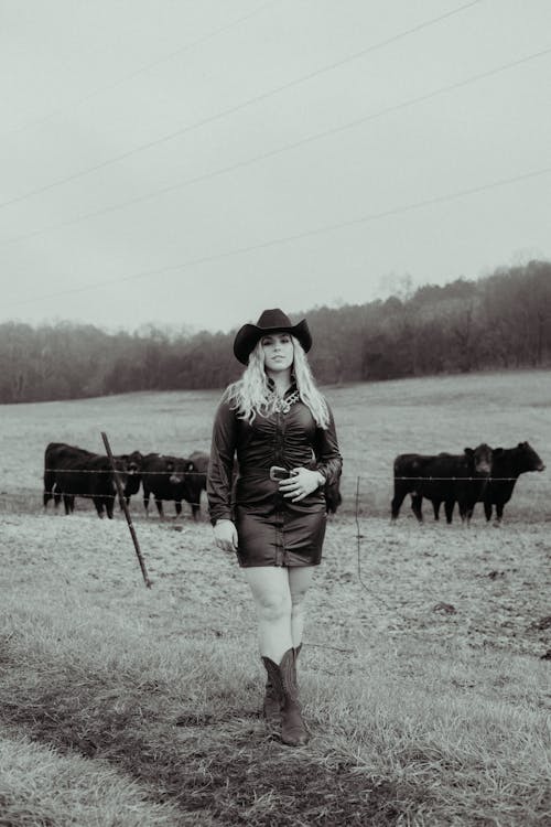 Blonde Woman in Black Dress Standing Near Cows on Pasture