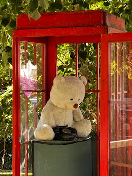 A cute teddy bear sits with an antique rotary phone inside a red phone booth in a sunny garden setting.