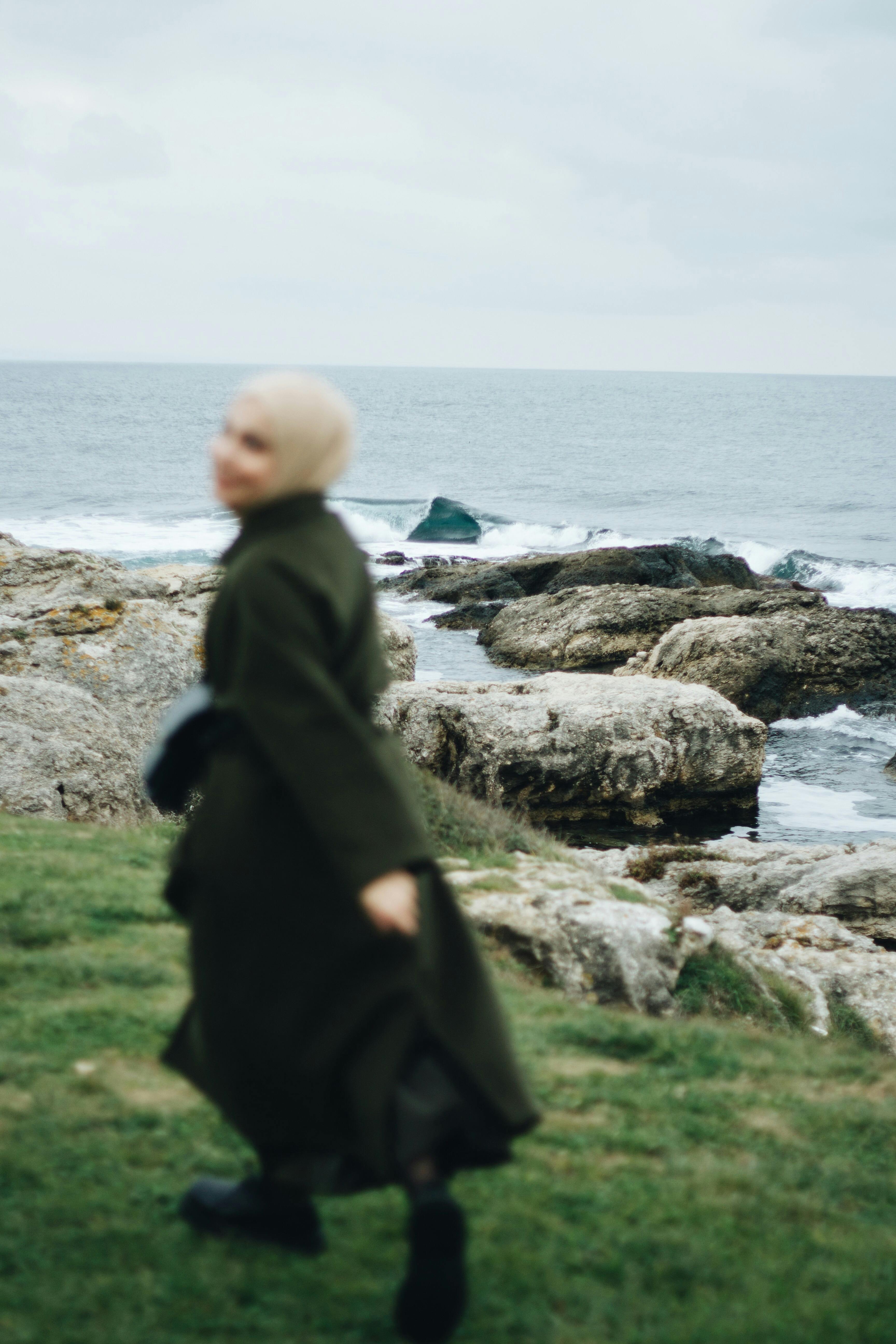 A blurred woman in a hijab walks along a rocky coastline, ocean waves in the background.