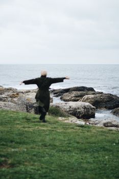 A woman joyfully runs with arms spread on a rocky shore by the sea, portraying freedom and nature's beauty.