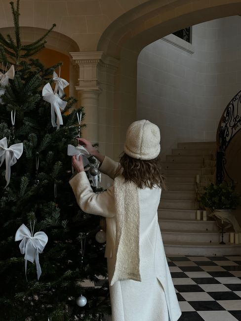 A woman in a cozy outfit decorates a Christmas tree with white bows in a stylish interior.