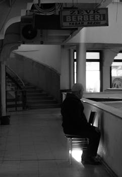 A man sits alone by the stairs in a quiet black and white setting.