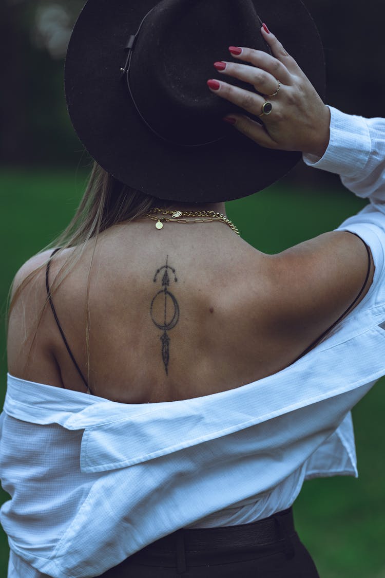 Back View Of A Woman With A Back Tattoo Standing Outside And Wearing A White Shirt And A Hat 