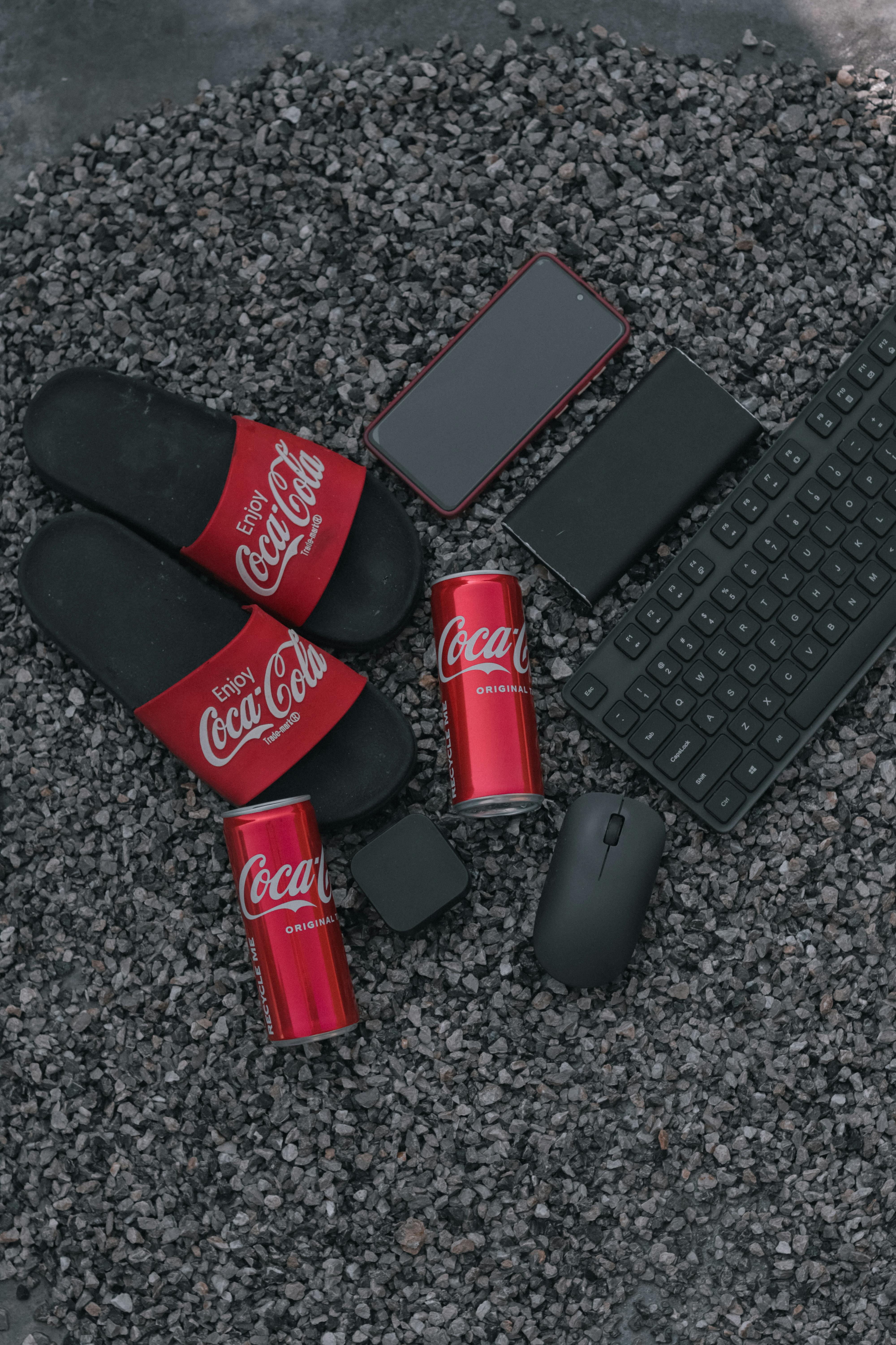 Flat lay with Coca Cola cans, smartphone, keyboard, and flip flops on gravel.