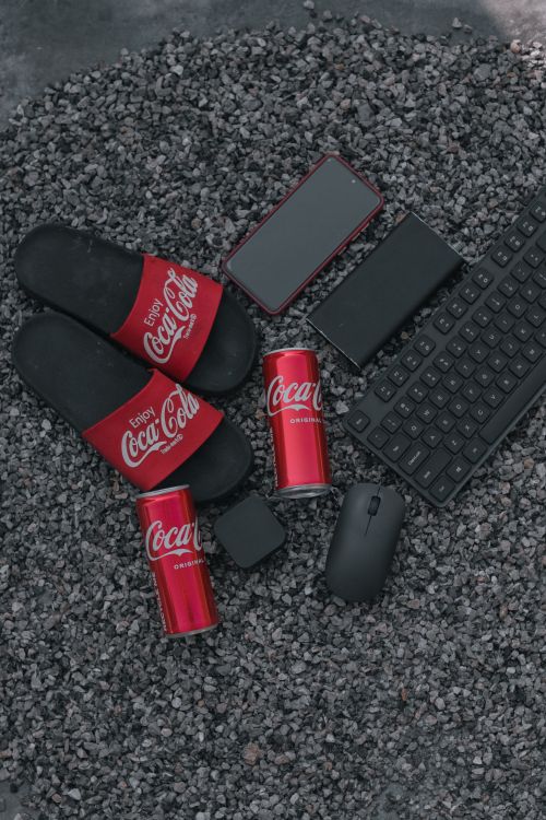 Free Flat lay with Coca Cola cans, smartphone, keyboard, and flip flops on gravel. Stock Photo