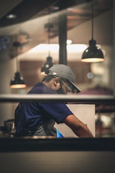 An Asian man in a cap and mask focused on work in a stylish cafe kitchen with warm lighting.