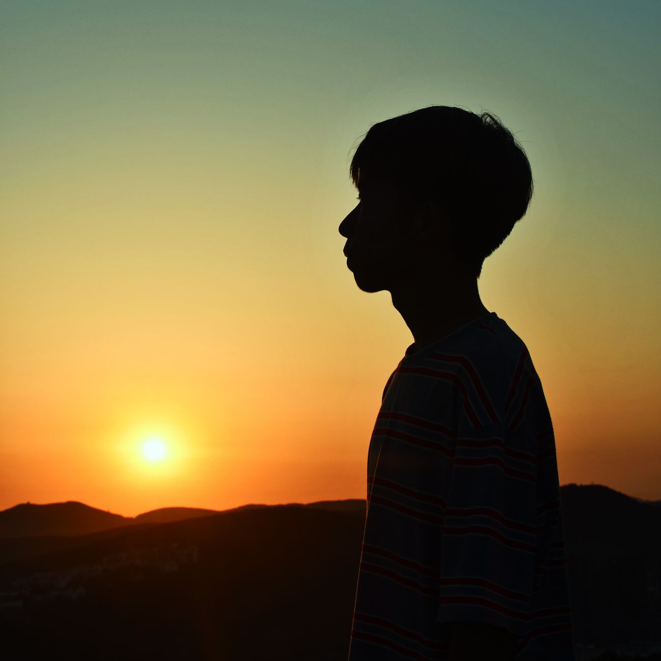 A serene silhouette of a boy watching a vibrant sunset over the hills, capturing a tranquil moment.
