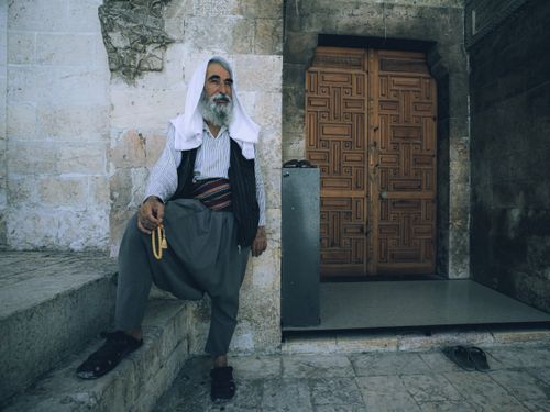 Free An elderly man wearing traditional clothing, sitting by a wooden doorway holding prayer beads. Stock Photo