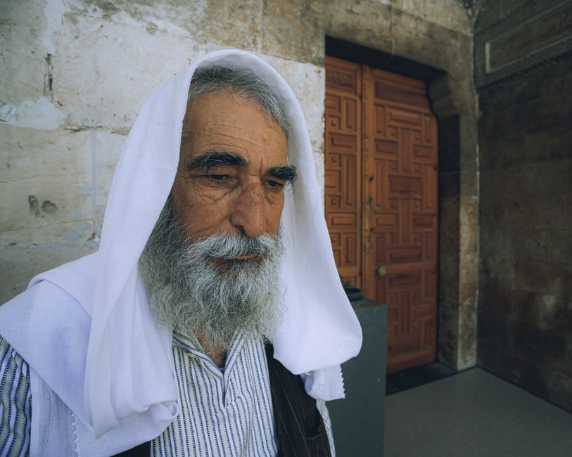 Senior man with white beard and hood stands by an ornate wooden door, exuding wisdom.