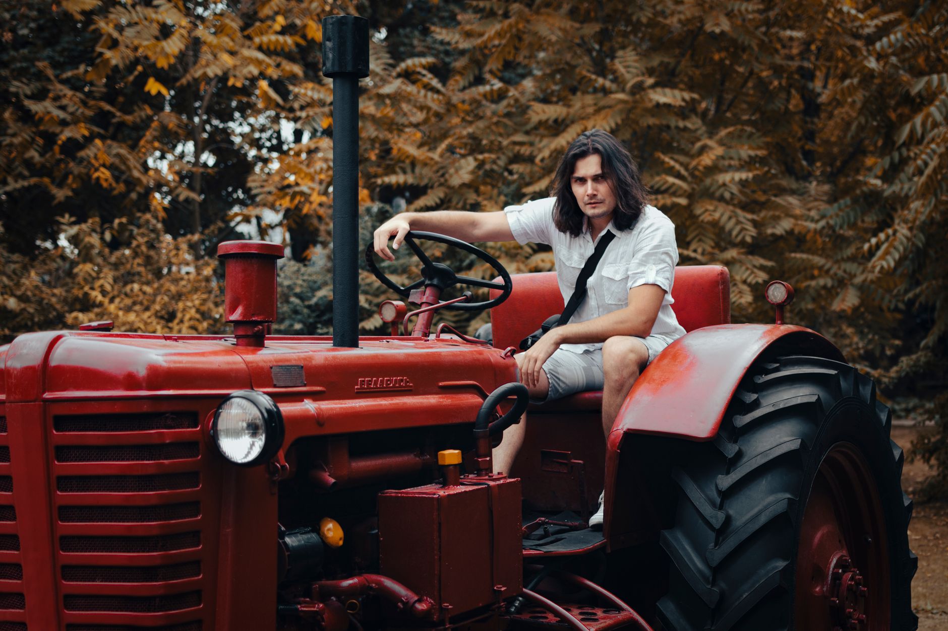 Man sitting on a red tractor in a forested outdoor setting, exuding a vintage rural vibe.