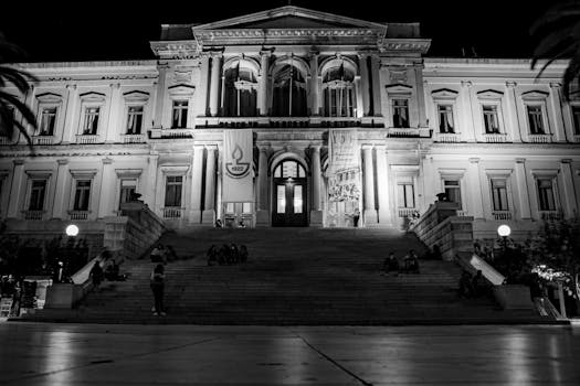 A black and white photo of a grand, illuminated historical building with symmetrical architecture taken at night.