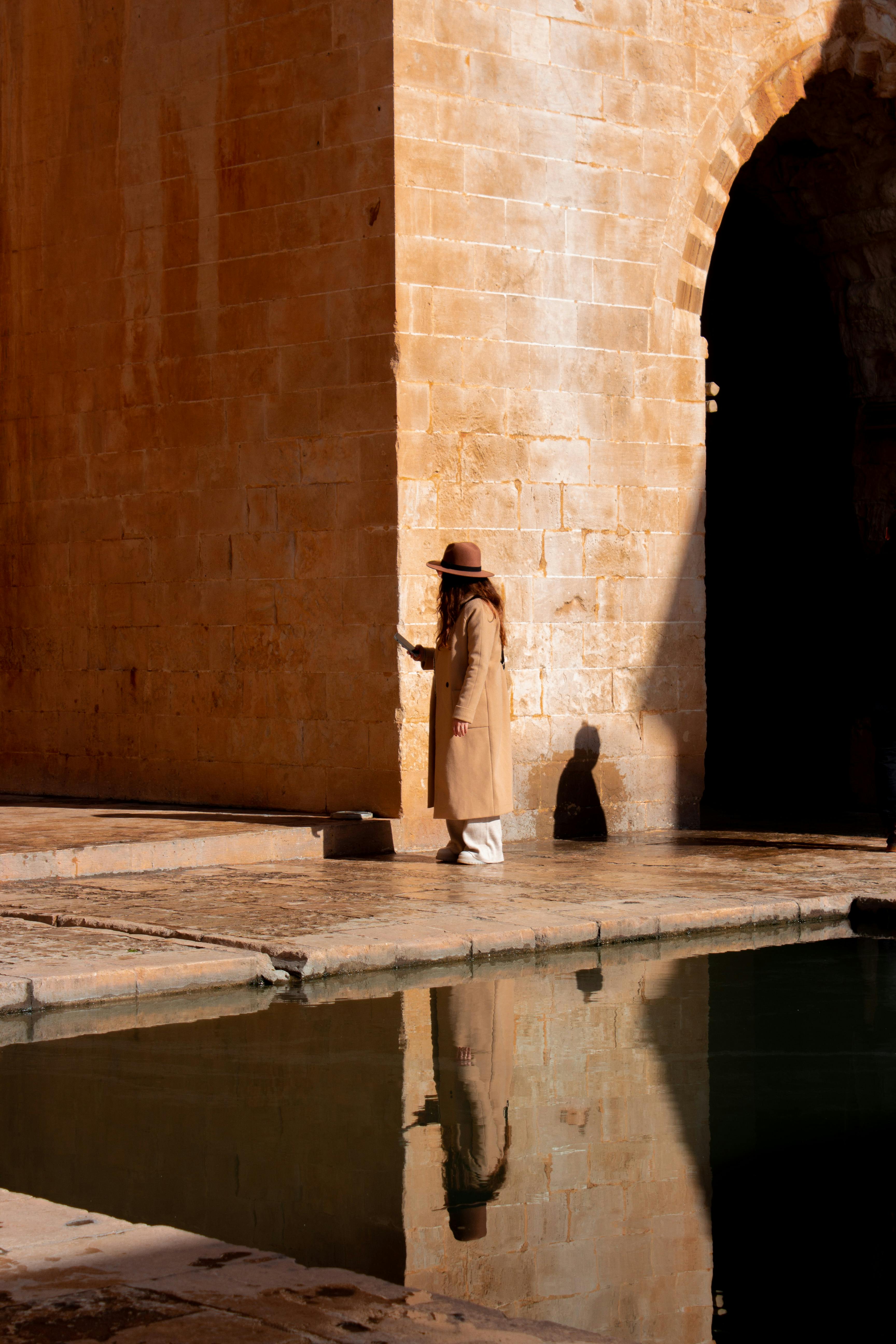 A woman in a brown coat stands by a historic stone archway with reflections in the water.