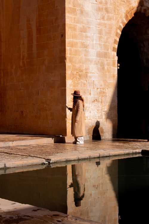 Free A woman in a brown coat stands by a historic stone archway with reflections in the water. Stock Photo