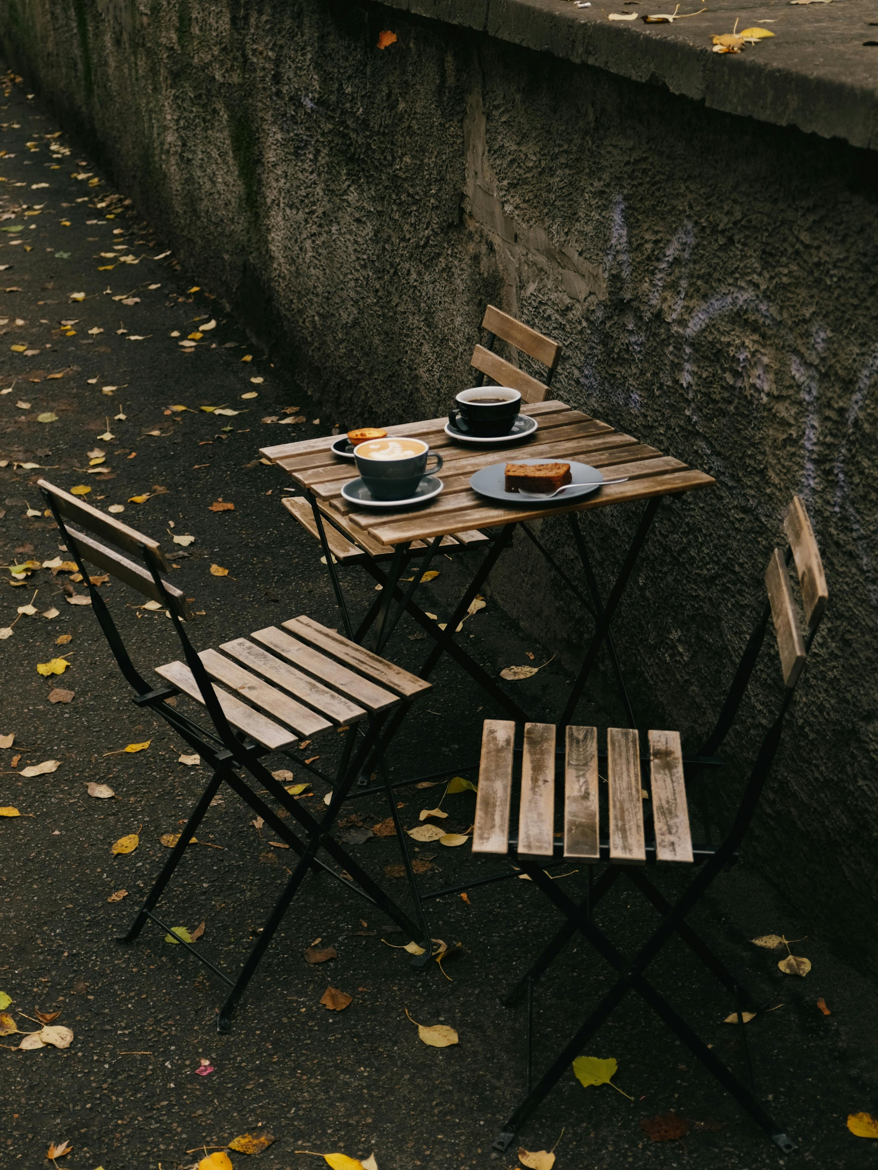 Charming outdoor cafe setting with dessert and coffee on a rustic table surrounded by fallen leaves.
