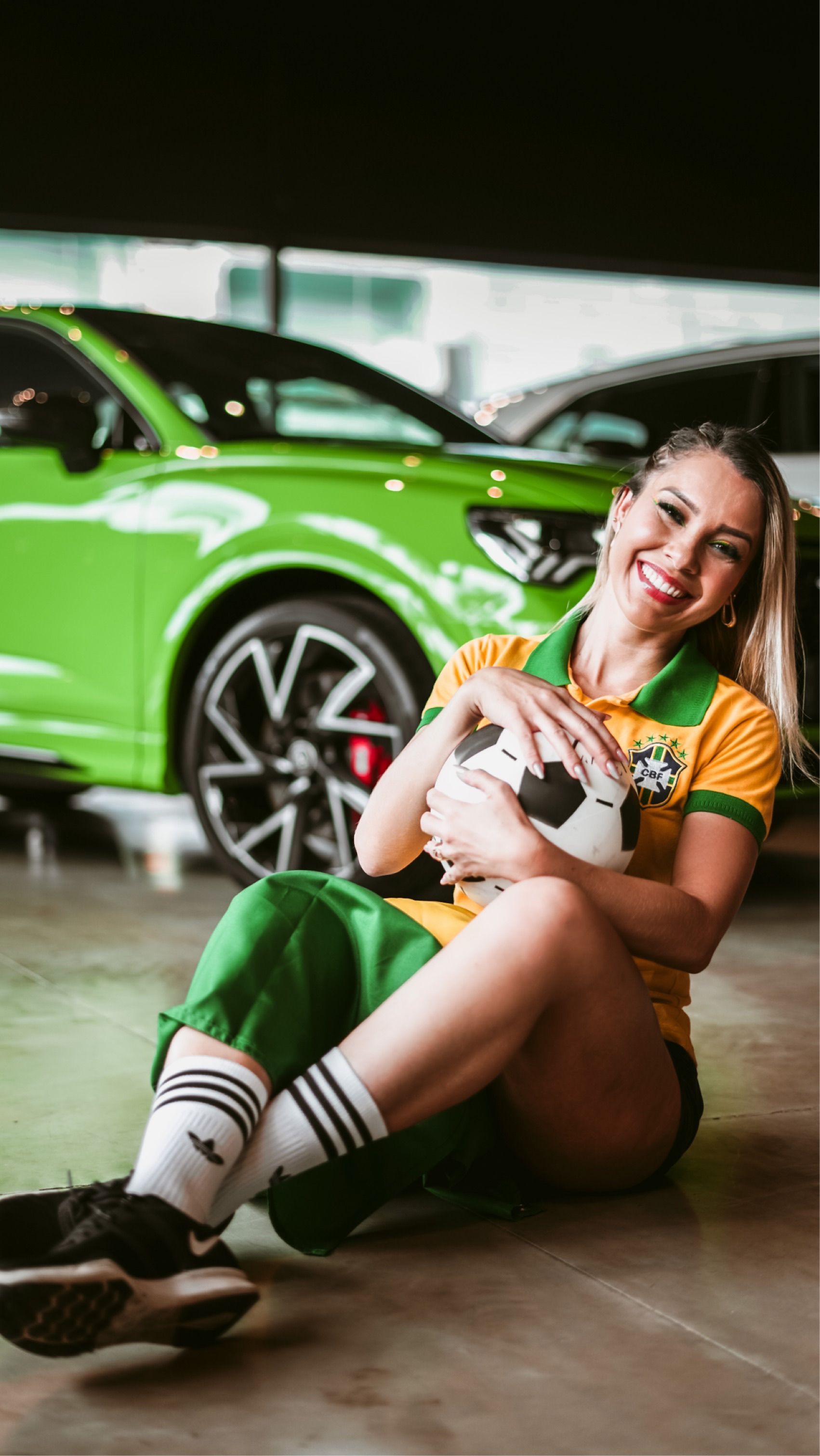 Smiling woman in sports attire hugging a soccer ball, sitting next to a vibrant green car indoors.