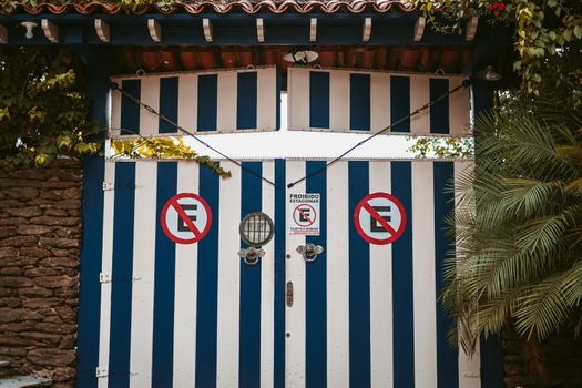 Blue and white striped gate with no parking signs amid lush greenery and stone walls.