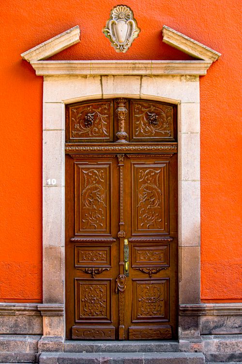Free A beautifully carved wooden door set against a vivid orange wall, exuding classic architectural elegance. Stock Photo