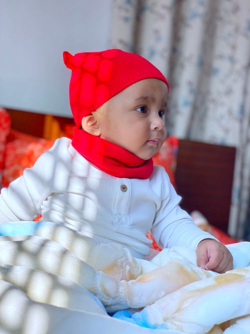 Free Charming baby wearing a red beanie and scarf, sitting indoors with soft natural light. Stock Photo