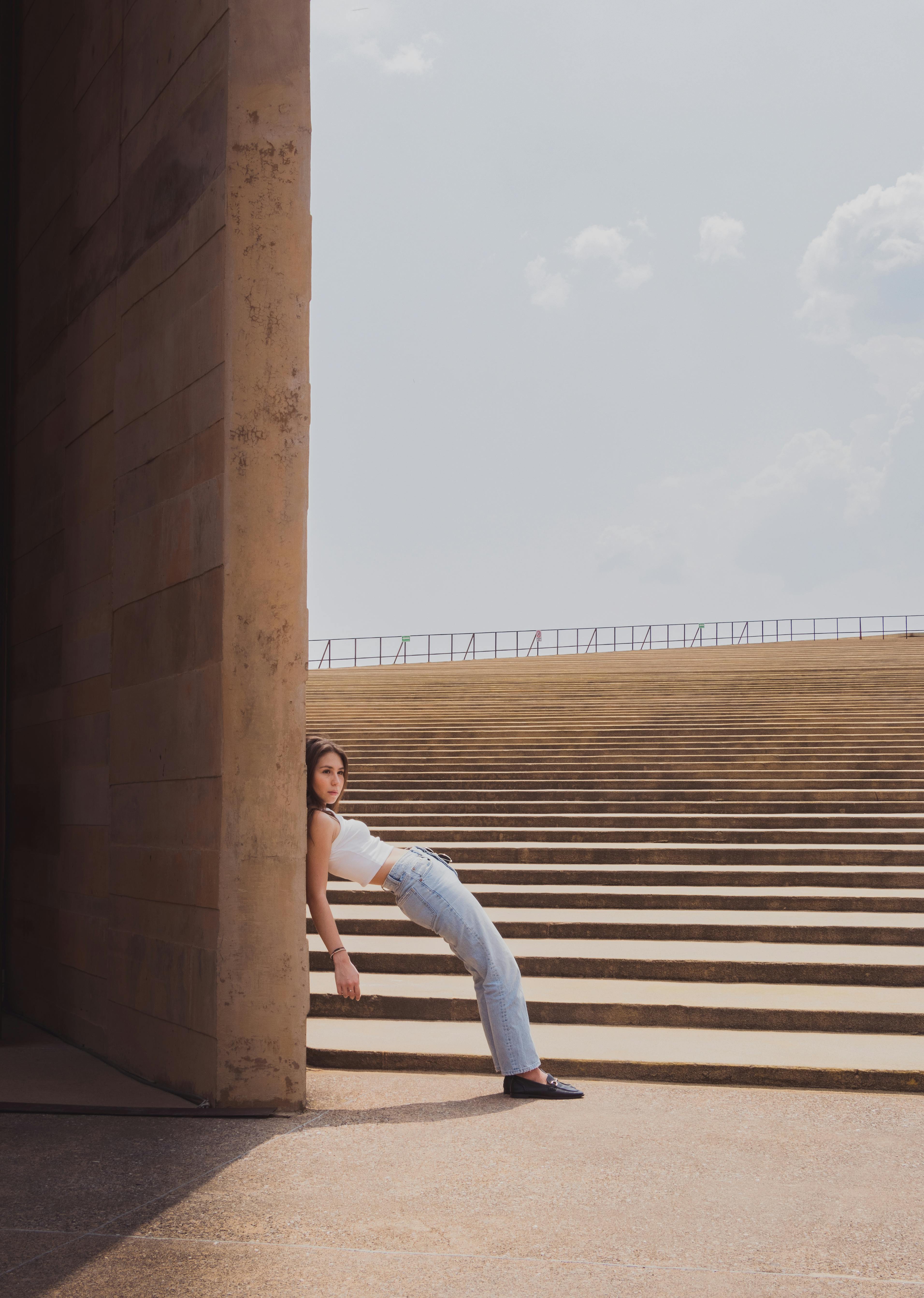 Stylish teenager in jeans and tank top leaning on stairs under a clear summer sky.