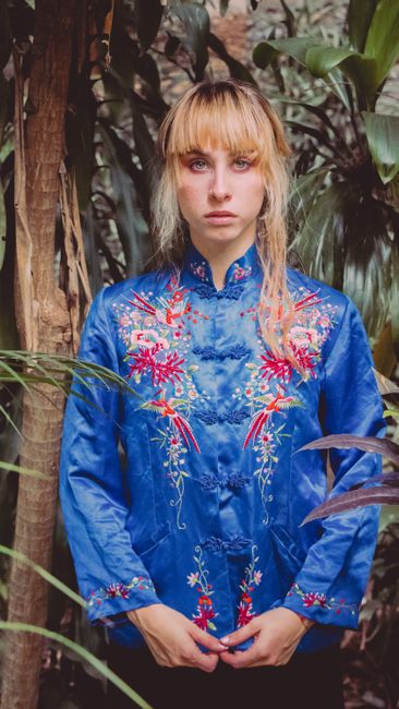 Portrait of a woman in an embroidered blouse standing amidst lush tropical foliage.