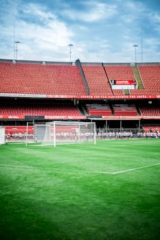 A vibrant empty soccer stadium with red seats and a goal post under a blue sky.