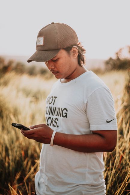 A young man in a cap texts on his smartphone in an outdoor field during the day.