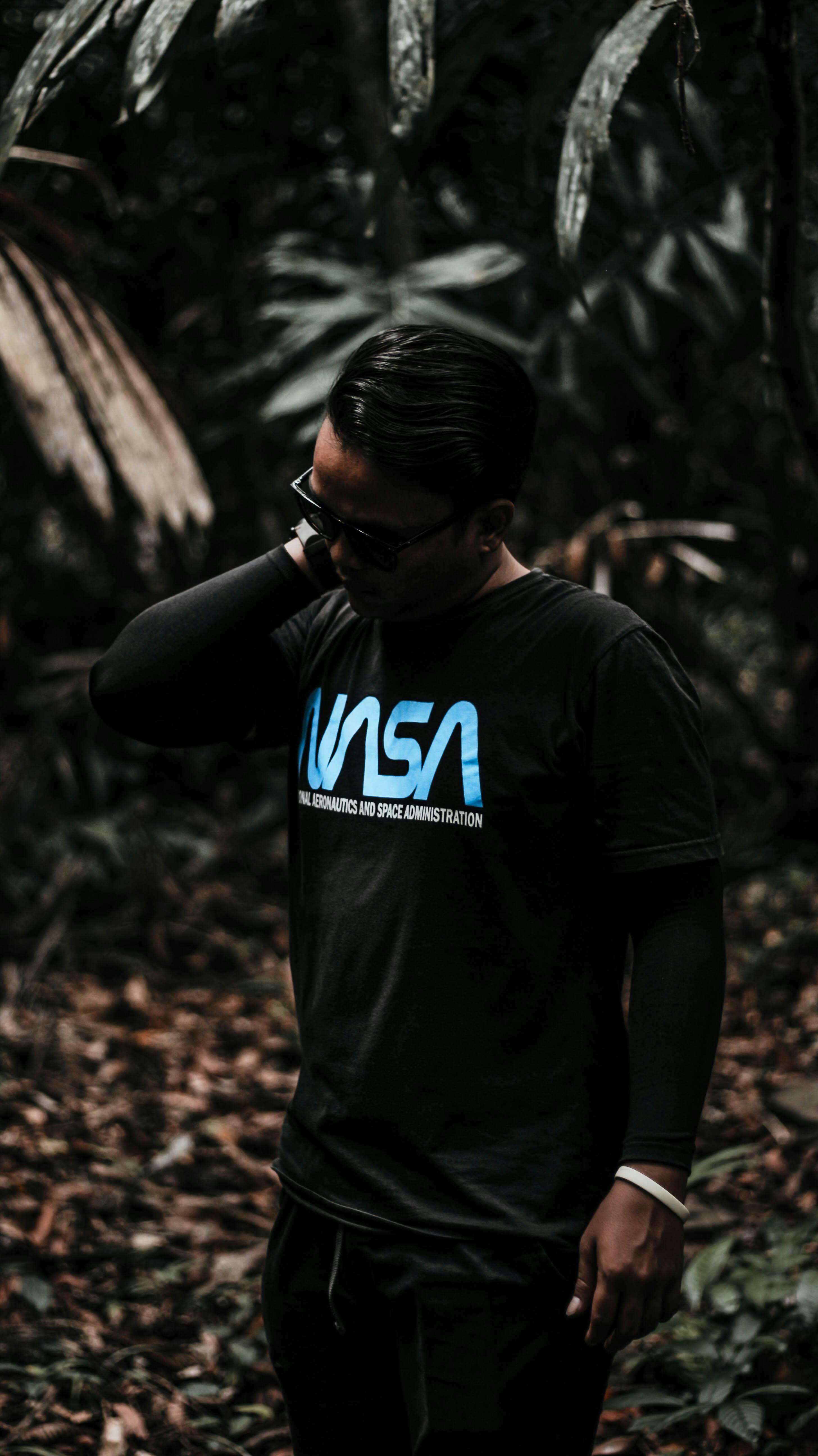 一个戴着眼镜、身着深色衣服的男人站在郁郁葱葱的森林里，营造出阴郁的氛围