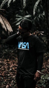 一个戴着眼镜、身着深色衣服的男人站在郁郁葱葱的森林里，营造出阴郁的氛围