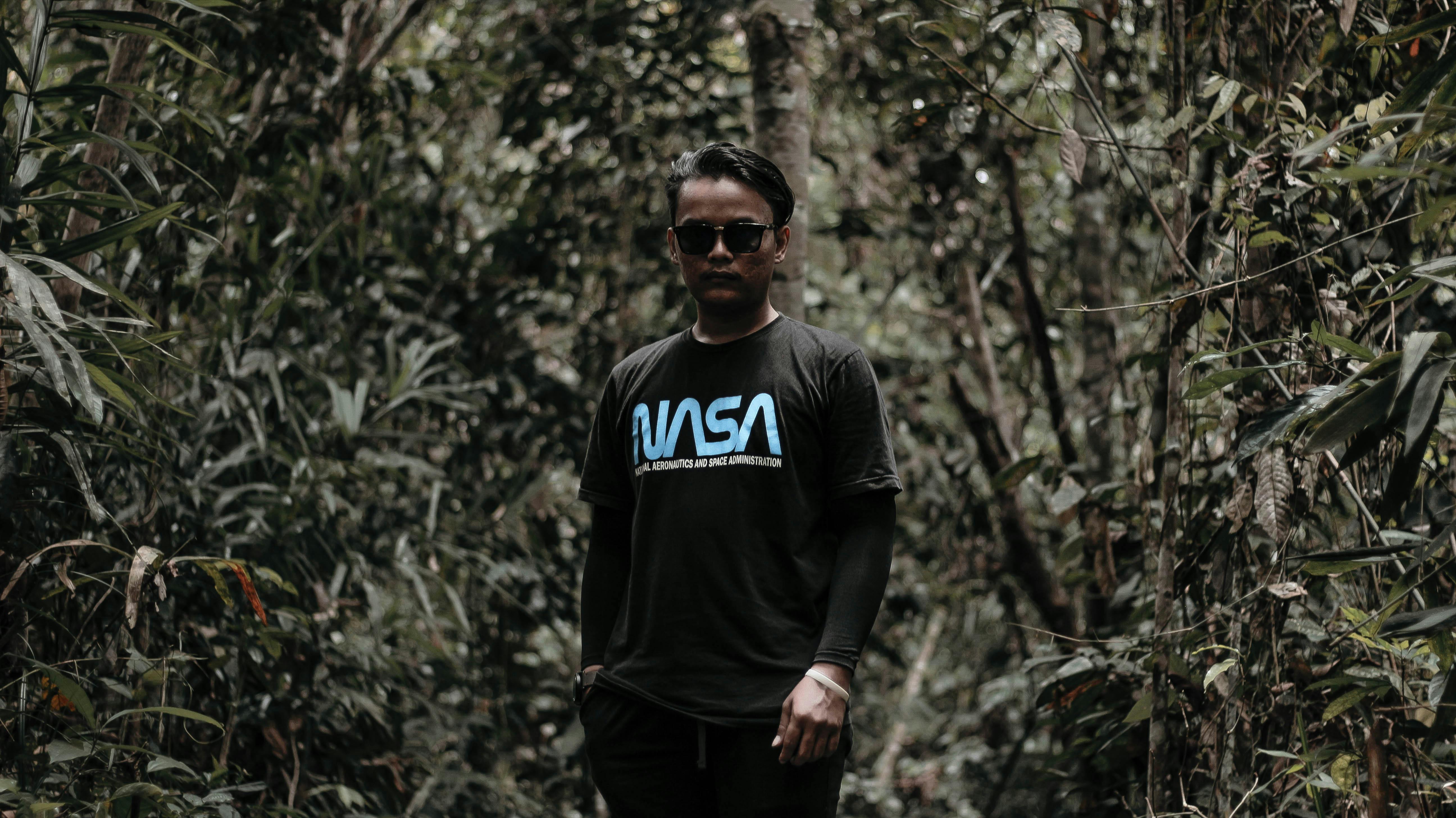 A man stands in a lush forest wearing a NASA t-shirt and sunglasses, surrounded by greenery.