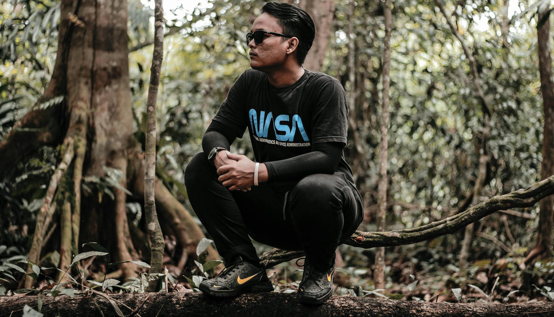 Man in sunglasses squatting on a tree branch wearing a stylish t-shirt in a forest setting.