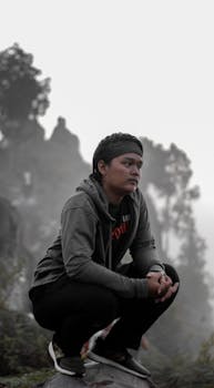 在朦胧幽静的森林中，一个人穿着连帽衫蹲在一块大岩石上，散发出平静而沉思的心情