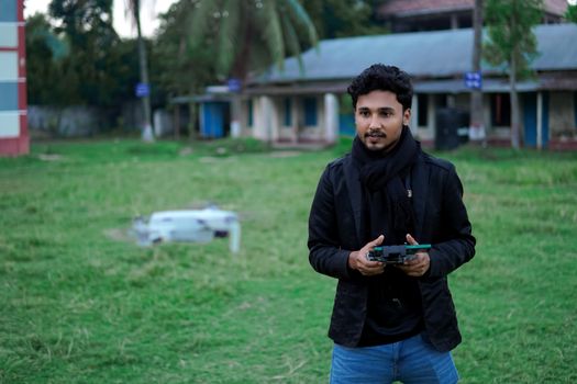 A young man in a jacket stands in a grassy field operating a drone controller.