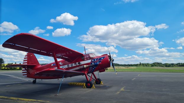 A historic red biplane parked on an airport tarmac under a vibrant blue sky with clouds.