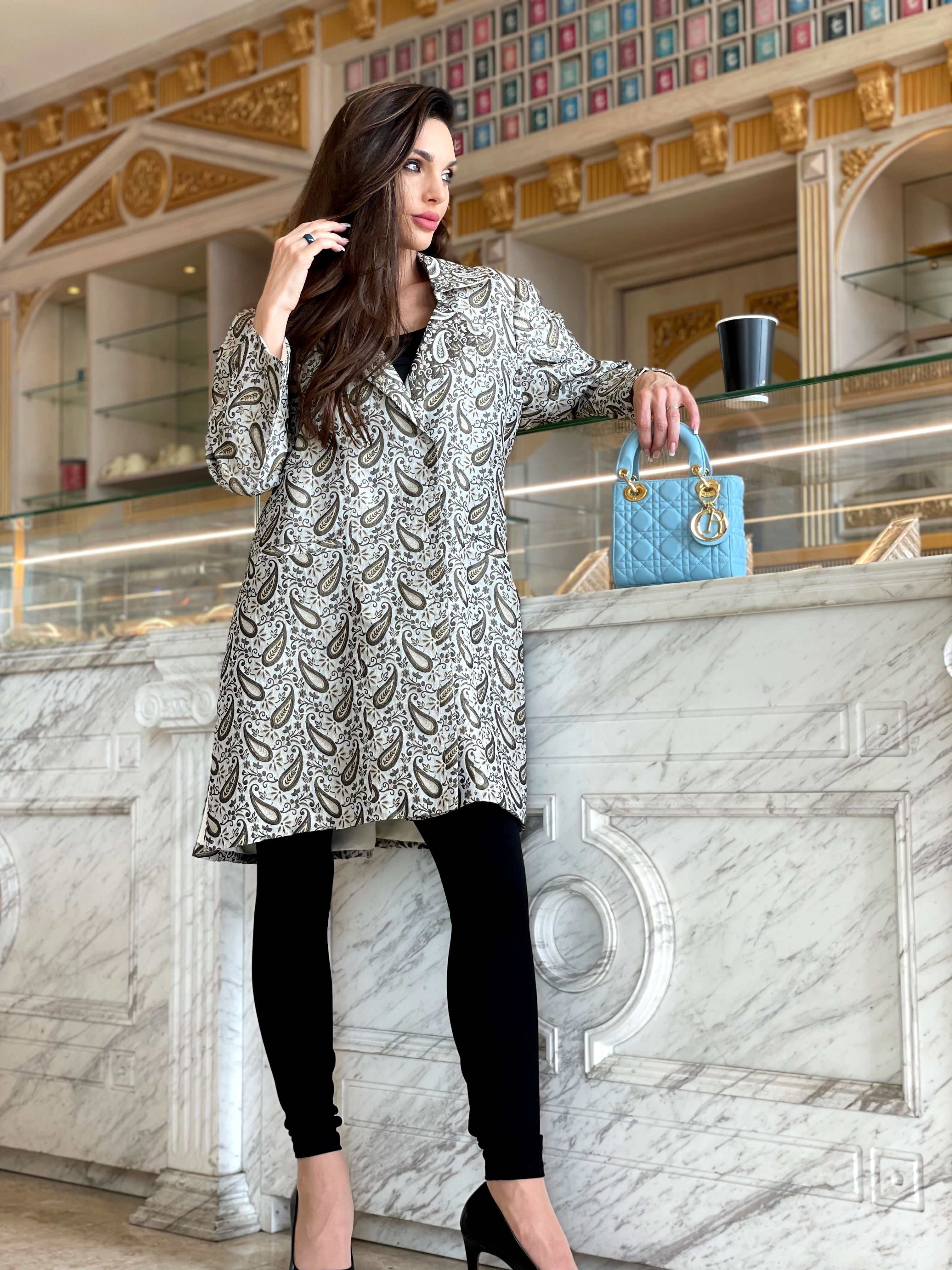 Modelo de moda chique em casaco estampado posando em um balcão de boutique luxuosa com uma bolsa azul e xícara de café.