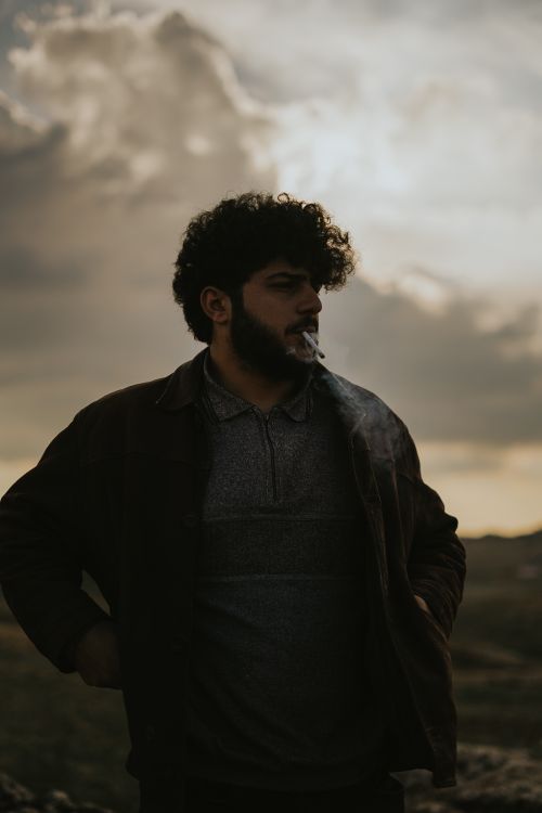 Free A young man with a beard and curly hair smokes outdoors under a dramatic sky. Stock Photo