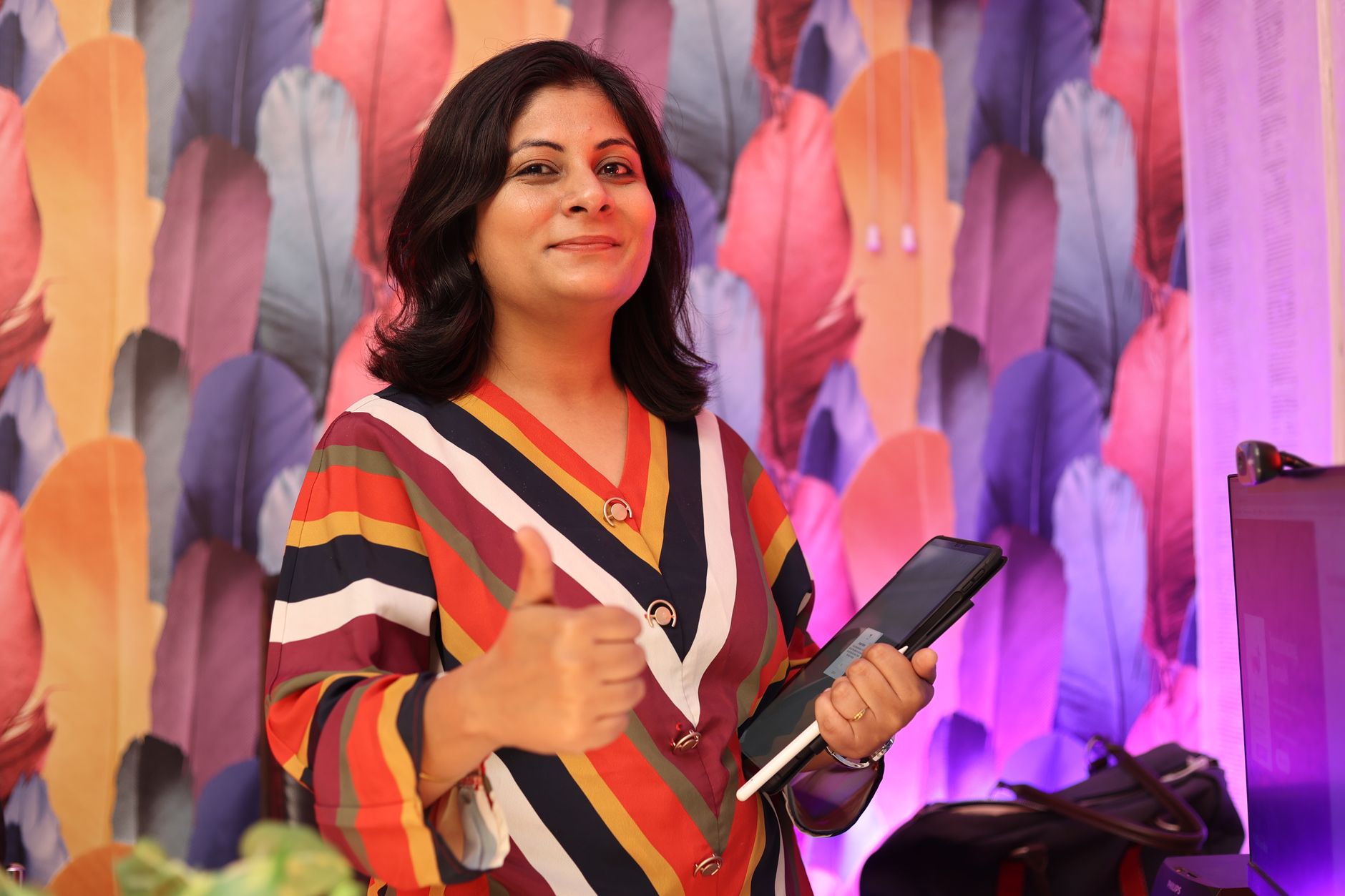 Smiling woman giving thumbs up while holding a tablet, standing indoors with a colorful background.