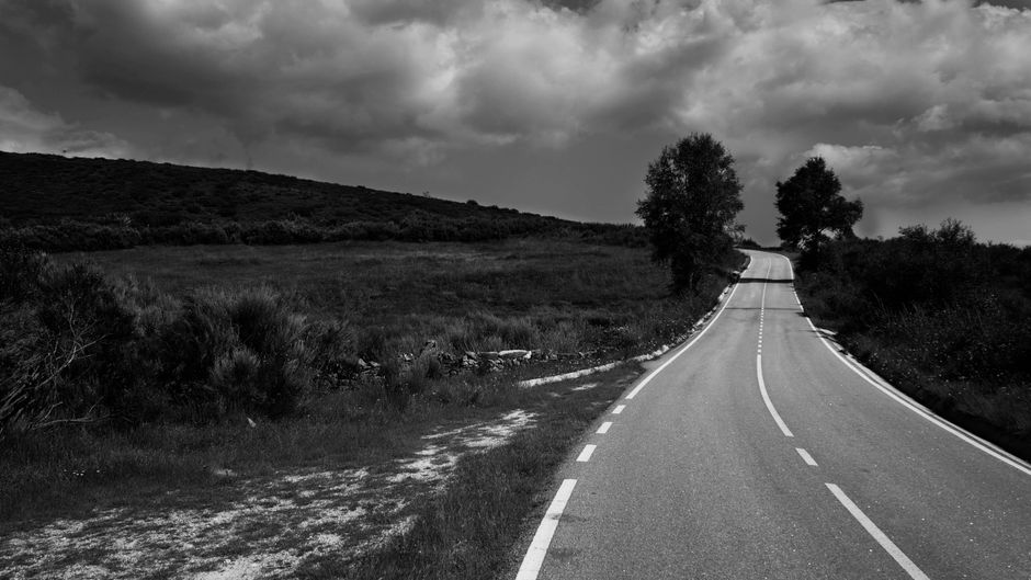 paved road, rolling hills, dramatic overcast, roadside greenery, travel atmosphere