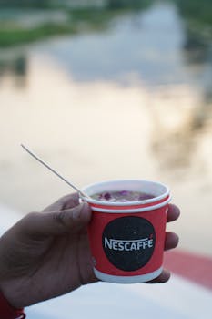 Hand holding a Nescafe disposable coffee cup with a serene lake in the background.